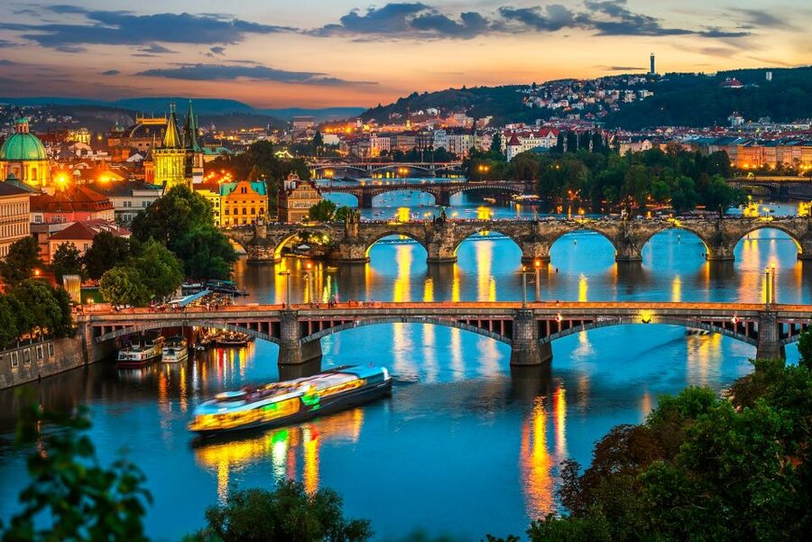 Lit bridges over the Vltava in Prague at dusk, a cruise boat mid-river